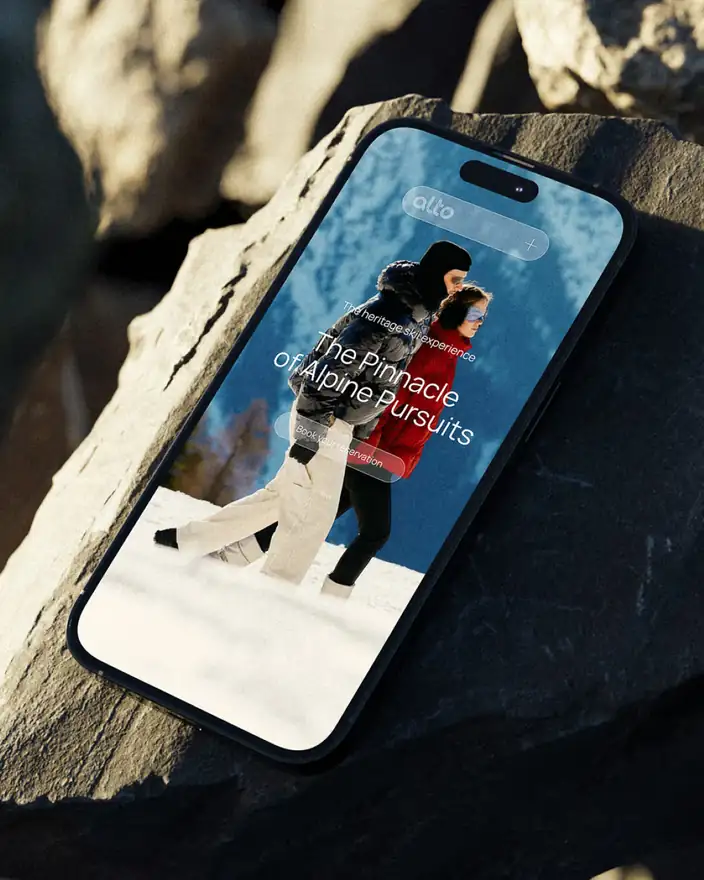 A phone on a rock showing the Alto mobile homepage with two skiers and a Book your reservation button