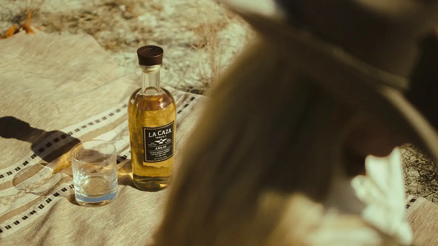 A bottle of La Caza A\u00f1ejo and a glass on a woven blanket in dry desert scrub at golden hour