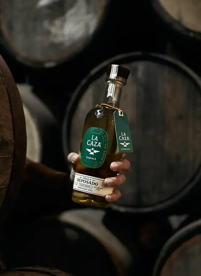 A hand holding La Caza Reposado in front of stacked ageing barrels