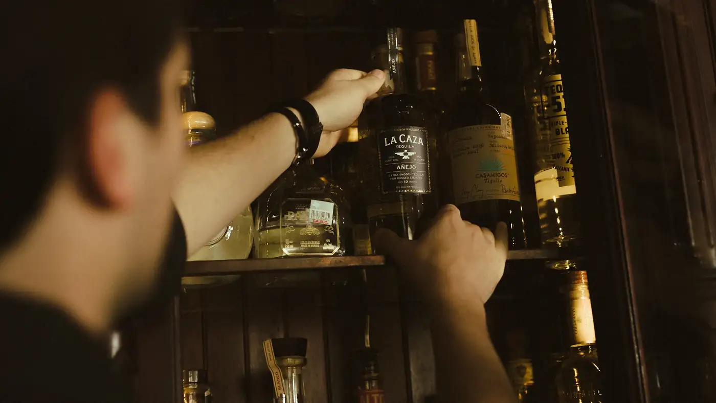 A bartender lifting a bottle of La Caza A\u00f1ejo from a crowded back-bar shelf