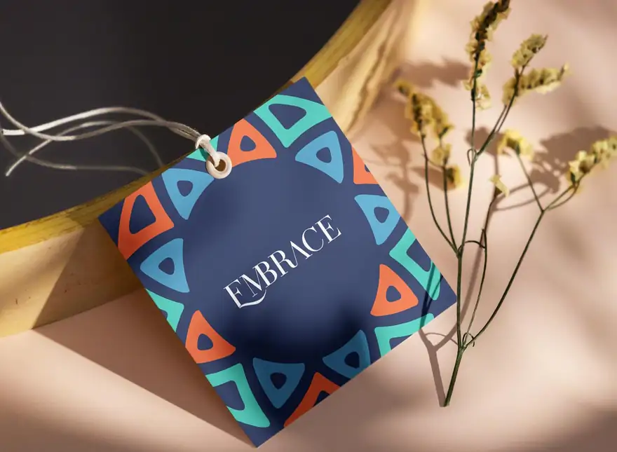 A square Embrace swing tag in navy, ringed with orange, teal and blue triangular motifs around the wordmark