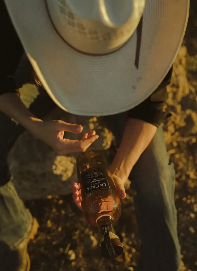 A rancher in a wide-brimmed straw hat holding a bottle of La Caza A\u00f1ejo, seen from above at golden hour